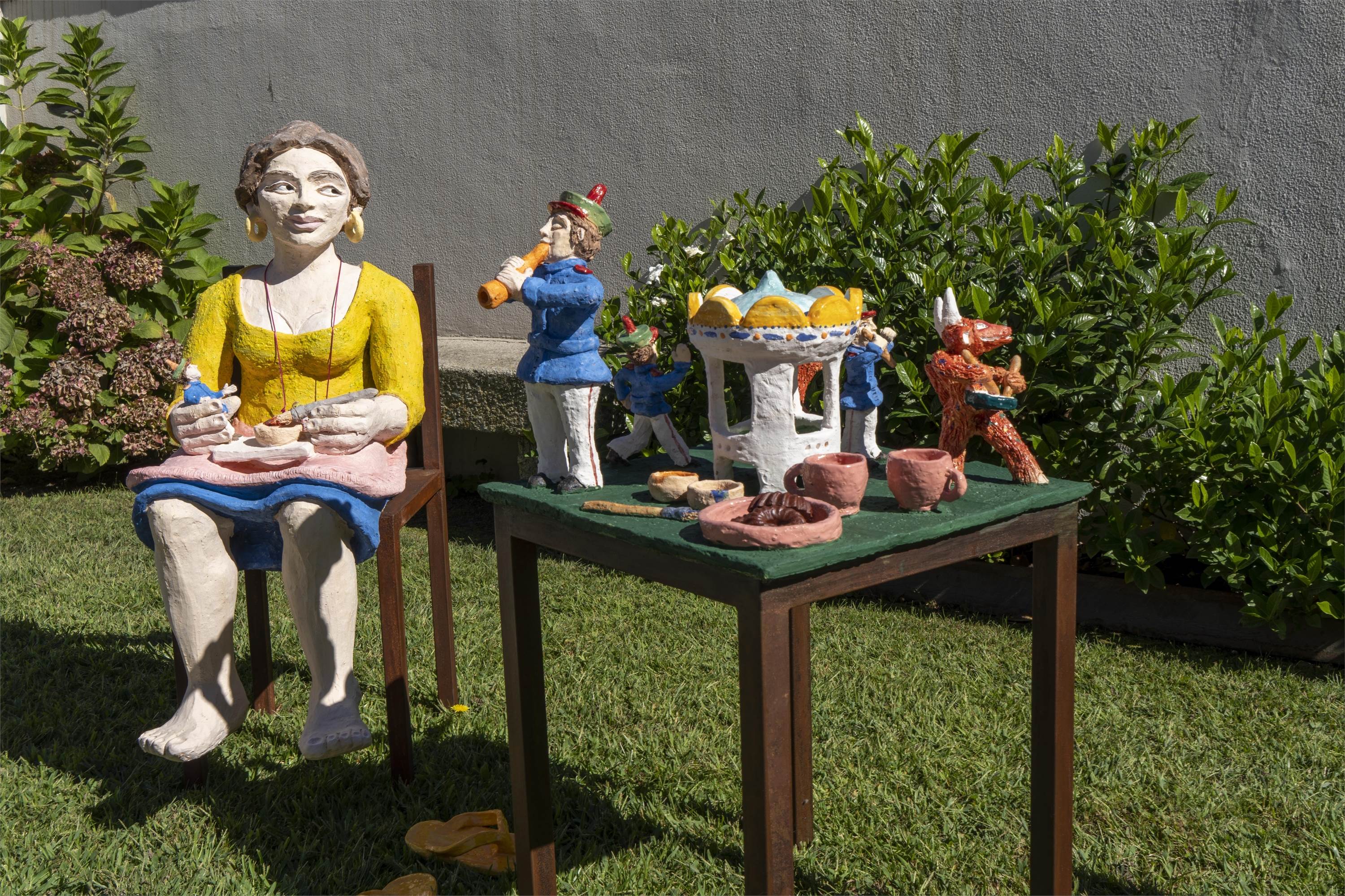 10.09.2022
Liliana Velho esteve em residência artística no Museu de Olaria de Barcelos. A intervenção artística retrata uma oleira em processo de trabalho, que constrói pequenos músicos para formar uma banda filarmónica, um dos vários temas do figurado de Barcelos. Enquanto trabalha é distraída pelo músico que toca flauta e não resiste a esboçar-lhe um sorriso. Na mesma mesa de trabalho outra ação decorre. A confusão instala-se com a invasão de pequenas criaturas travessas, os Diabos, que se divertem roubando os instrumentos aos músicos e fazendo-os perder a compostura. O cenário é uma espécie de realismo mágico onde se imaginam histórias entre as diferentes figuras singulares do figurado de Barcelos, um entrelaçar da realidade com a fantasia, tentando retratar uma pequena parte do ofício da cerâmica e homenagear os artesãos locais, as temáticas e tradições de Barcelos.