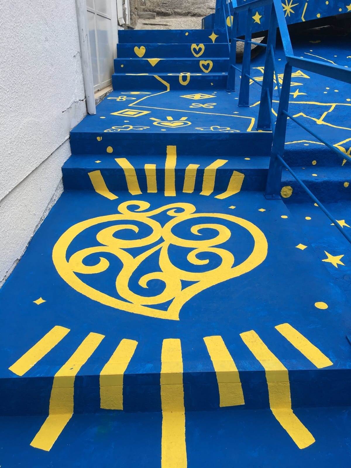 25.04.2022
The people of Póvoa do Lanhoso inherited from the sharpness of the slab, which marks its geography, the strength and perseverance that we also recognise in the personalities that are the heritage of the land. The symbolism of the slab lies in the courage of Maria da Fonte as in the talent of its goldsmiths and craftsmen. The challenge set to Mariana Mizarela was to intervene in a neighbour’s staircase that unites two worlds in that place. The artist worked with the colours of the Póvoa do Lanhoso flag which, coincidentally, are also the colours of the Ukrainian flag and on the 25th of April 2022, with Europe at war, we also celebrate the importance of Freedom. With gold on blue a constellation is drawn, a sky on the ground, which unites the various symbols of that place. The intervention went beyond the sinuosity of the staircase and its execution deserved the collaboration of the local community that now has a new place for meeting and peace.